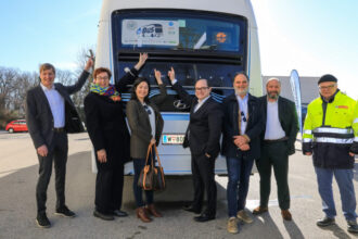 Personen stehen vor der Rückseite des Busses und zeigen auf Logo des Projektes