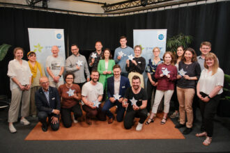 Eine Gruppe von 17 Personen posiert gemeinsam vor zwei „greenstart“-Bannern, einer Start-up-Initiative für Klima- und Energielösungen. Viele von ihnen halten sternförmige Schilder mit ihren Unternehmensnamen und lächeln stolz in die Kamera. Die Atmosphäre wirkt professionell und zugleich freundlich, mit einer Mischung aus Business- und Casual-Kleidung.
