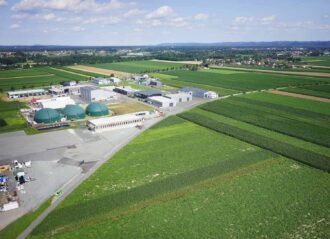 Luftaufnahme einer modernen Biogasanlage inmitten landwirtschaftlicher Felder. Mehrere große grüne Kuppeln sind sichtbar, umgeben von Industriegebäuden und befestigten Flächen. Im Hintergrund erstrecken sich Felder, kleine Ortschaften und eine hügelige Landschaft unter einem leicht bewölkten Himmel.