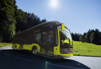 Von schräg vorne ein elektrischer gelber Bus, im Hintergrund blauer Himmel, eine Wiese und Bäume.