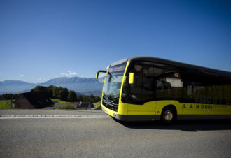 Von der Seite das Vorderteil eines gelben elektrischen Busses, im Hintergrund blauer Himmel und Berge.