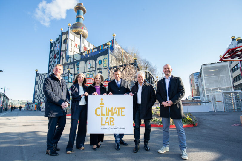 Eine Gruppe von sechs Personen hält ein Schild mit der Aufschrift 'Climate Lab' und der Website www.climatelab.at. Im Hintergrund ist die bunte, ikonische Architektur eines Gebäudes mit goldenen Kugeln und Schornsteinen zu sehen, das bei sonnigem Wetter fotografiert wurde.