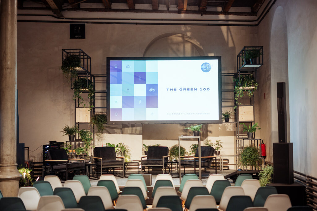 Eine modern gestaltete Eventbühne für „The Green 100 – Die grüne Finanzierungsmessen“, mit einer großen Leinwand, auf der das Logo des Klima- und Energiefonds zu sehen ist. Die Bühne ist mit schwarzen Sesseln, kleinen Tischen und zahlreichen Pflanzen dekoriert, um eine nachhaltige Atmosphäre zu schaffen. Im Vordergrund sind Stuhlreihen in Grün- und Weißtönen für das Publikum angeordnet.