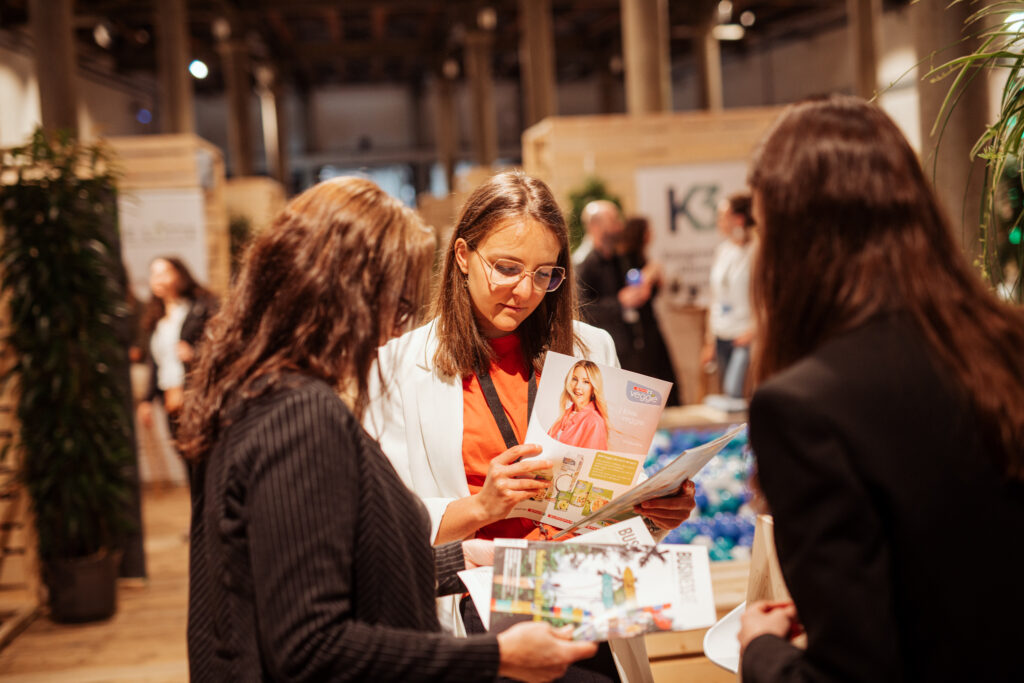 Drei Frauen in Business-Kleidung stehen in einem modernen, offenen Messebereich und betrachten Broschüren zu nachhaltigen Themen. Eine Frau im weißen Blazer mit rotem Oberteil hält eine Broschüre mit dem Titel „I love veggie“ in der Hand. Im Hintergrund sind weitere Messestände, Pflanzen und ein mit blauen und weißen Bällen gefülltes Becken zu sehen.