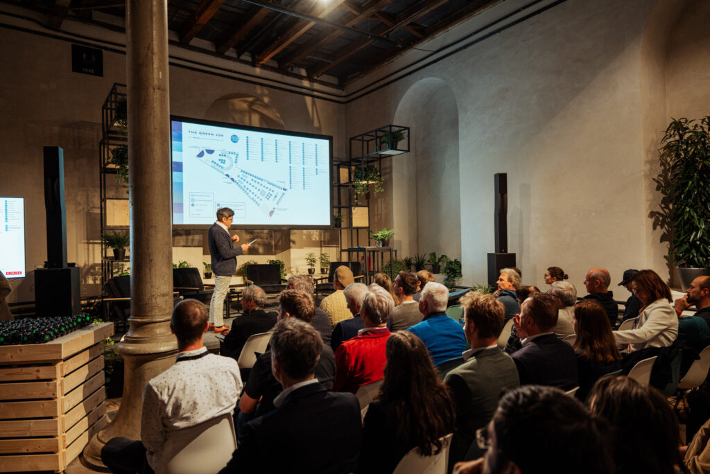 Ein Sprecher steht vor einem Publikum in einem historischen Saal mit hohen Decken und präsentiert eine Karte der Veranstaltung „The Green 100“ auf einer großen Leinwand. Die Zuhörer, viele von ihnen in Business-Kleidung, verfolgen aufmerksam die Präsentation. Die Bühne ist mit Pflanzen dekoriert, und das Ambiente wirkt modern und nachhaltig.