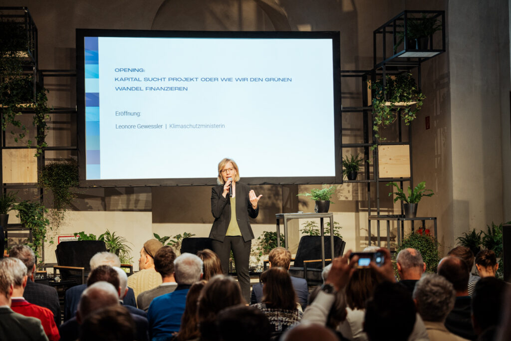 Eine Rednerin steht auf einer Bühne vor einem aufmerksamen Publikum und hält eine Präsentation zum Thema „Kapital sucht Projekt oder wie wir den grünen Wandel finanzieren.“ Die Leinwand zeigt den Namen der Klimaschutzministerin Leonore Gewessler als Eröffnungsrednerin. Die Bühne ist mit modernen Metallregalen und Pflanzen dekoriert, was eine nachhaltige und inspirierende Atmosphäre schafft.