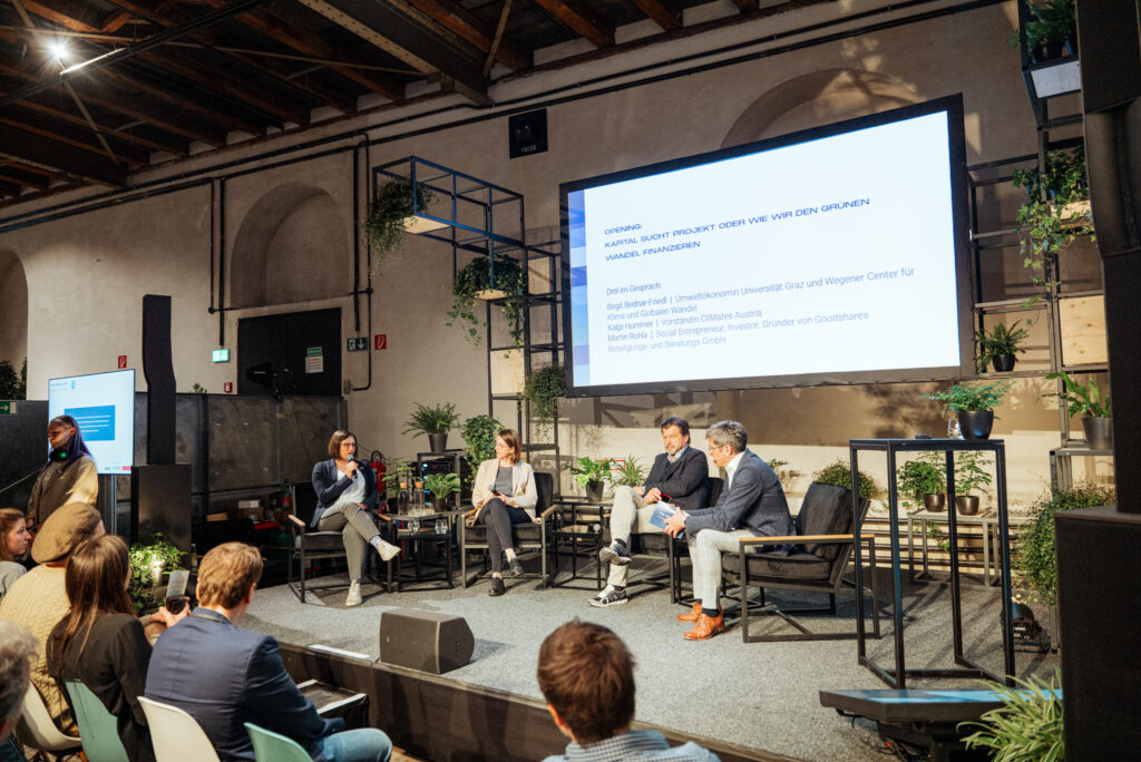 Eine Podiumsdiskussion auf einer Konferenzbühne mit vier Panelisten, die über nachhaltige Finanzierung sprechen. Eine Frau mit Mikrofon stellt eine Frage, während die anderen Teilnehmer aufmerksam zuhören. Im Hintergrund zeigt eine große Leinwand den Titel der Diskussion: „Kapital sucht Projekt oder wie wir den grünen Wandel finanzieren.“ Das Setting ist modern gestaltet, mit vielen Pflanzen und einer offenen Raumarchitektur.