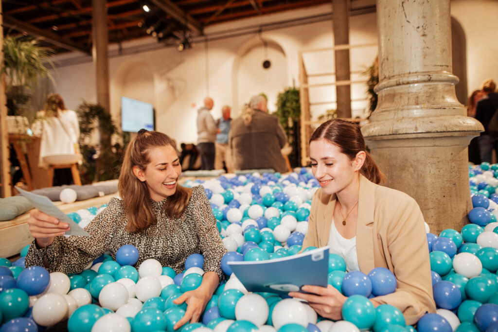 Zwei junge Frauen sitzen in einem Ballpit aus blauen, weißen und türkisfarbenen Kugeln und tauschen sich fröhlich über Dokumente aus. Eine Frau in einer beigen Blazerjacke hält eine blaue Mappe, während die andere in einem gemusterten Oberteil ein Blatt Papier hochhält. Im Hintergrund sind weitere Messebesucher und ein offener, moderner Veranstaltungsraum mit Pflanzen und Sitzgelegenheiten zu sehen.