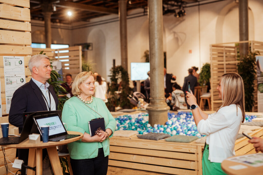 Eine Frau in einem weißen Blazer macht mit ihrem Smartphone ein Foto von zwei Personen, die für die Kamera posieren. Die Szene spielt sich in einer modernen Messe- oder Networking-Umgebung ab, mit Holztrennwänden, Informationsmaterial und einem bunten Bällebad als kreatives Element. Im Vordergrund stehen ein Mann im Anzug und eine Frau in einem mintgrünen Oberteil mit einer Perlenkette, beide wirken professionell und freundlich.