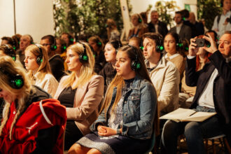 Eine Gruppe von Menschen sitzt konzentriert in einer Konferenz oder Präsentation, alle tragen schwarze Kopfhörer mit leuchtend grünen Lichtringen. Die Teilnehmer sind in Business- und Casual-Kleidung gekleidet, einige machen sich Notizen, während andere aufmerksam zuhören. Im Hintergrund steht ein Mann mit Smartphone und filmt oder fotografiert die Veranstaltung.