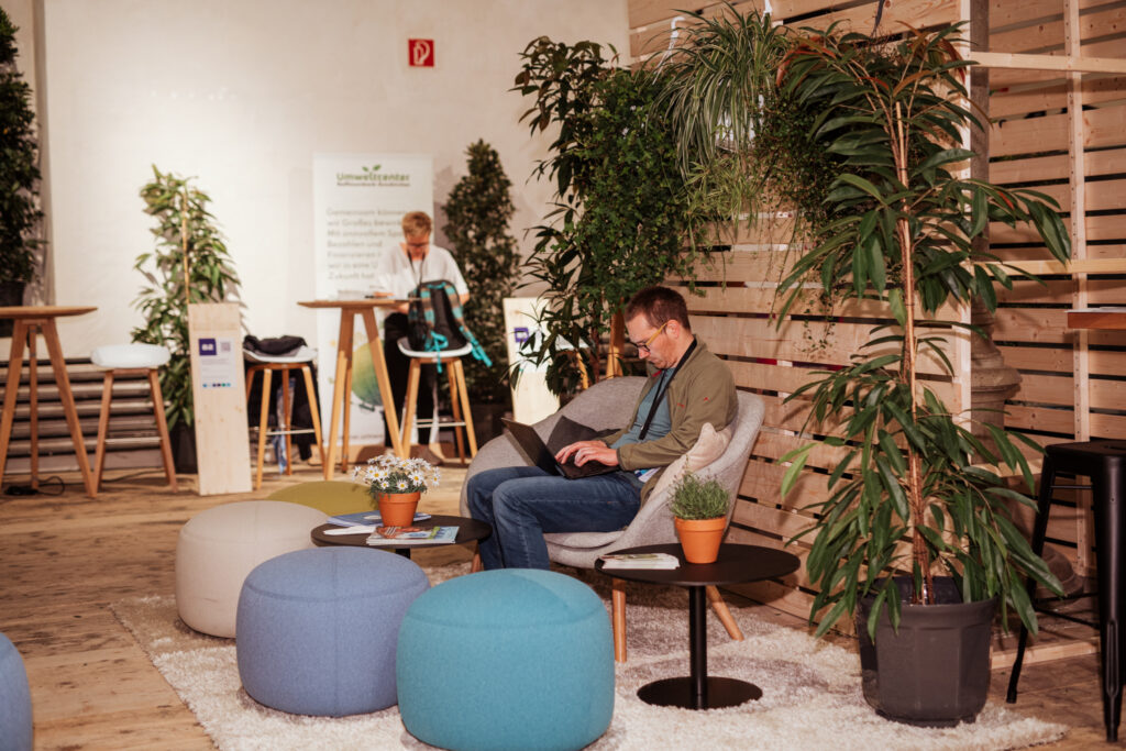Ein gemütlicher Lounge-Bereich mit runden, gepolsterten Sitzhockern in Blau- und Grüntönen sowie einem modernen Sessel, in dem ein Mann mit Laptop arbeitet. Umgeben von zahlreichen Pflanzen und Holztrennwänden strahlt die Umgebung eine entspannte Atmosphäre aus. Im Hintergrund sitzt eine weitere Person an einem Stehtisch mit Barhockern, während ein Banner mit dem Schriftzug 