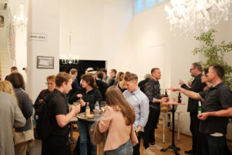 Eine lebhafte Networking-Veranstaltung in einem stilvoll beleuchteten Raum mit Kronleuchtern. Gäste stehen in kleinen Gruppen, unterhalten sich und genießen Getränke an Stehtischen. Ein Schild an einer Säule weist auf eine „Sharing Area“ hin, was auf einen kollaborativen oder nachhaltigen Event-Kontext hindeutet.