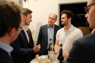 Eine Gruppe von Männern steht um einen Stehtisch bei einer Networking-Veranstaltung und unterhält sich angeregt. Leere Gläser und Bierflaschen auf dem Tisch deuten auf eine entspannte Atmosphäre hin. Die Teilnehmer tragen Business-Casual-Kleidung und wirken fokussiert auf das Gespräch.