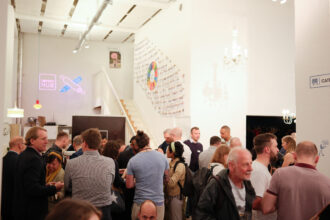 Eine Networking-Veranstaltung in einem modernen Raum mit hohen Decken, weißen Wänden und Kronleuchtern. Viele Menschen stehen in kleinen Gruppen, unterhalten sich und genießen Getränke. Ein Neon-Schild mit der Aufschrift „Impact Hub“ ist an der Wand zu sehen, während eine Treppe zu einem oberen Bereich führt.