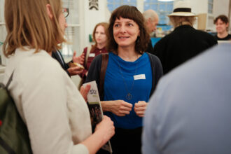 Eine Frau mit schulterlangem braunen Haar und blauer Kleidung lächelt während eines Gesprächs bei einem Networking-Event. Sie trägt ein Namensschild und eine lange Kette, während sie mit einer anderen Person spricht, die eine Broschüre hält. Im Hintergrund unterhalten sich weitere Gäste, einige mit Snacks und Getränken in der Hand.
