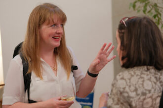 Eine Frau mit langen, rötlichen Haaren und einem Namensschild spricht lächelnd mit einer anderen Person bei einem Networking-Event. Sie trägt eine weiße Bluse, eine goldene Halskette und hält ein Weinglas in der Hand, während sie gestikulierend erzählt. Die Gesprächspartnerin, die eine Brille auf dem Kopf trägt, hört aufmerksam zu.