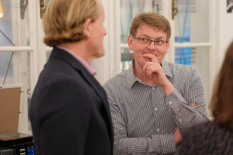 Ein Mann mit Brille und kariertem Hemd lehnt nachdenklich seinen Kopf auf die Hand, während er einem Gesprächspartner in einem dunklen Sakko zuhört. Im Hintergrund sind große Fenster mit weißen Rahmen und dekorativen Schnüren zu sehen, die eine gemütliche Atmosphäre schaffen. Der Ausdruck des Mannes vermittelt Interesse und Nachdenklichkeit in der Gesprächssituation.