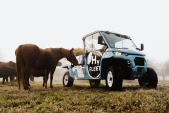 Ein blaues Nutzfahrzeug mit dem Schriftzug HyFleet steht auf dem Feld neben einem braunen Pferd.