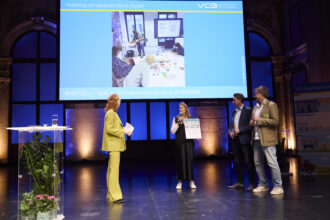 Bühne mit 4 Personen, eine Person hält einen Preis und ein Mikrofon in der Hand