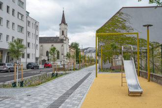 Städtische Straße, junge Bäume, Straße mit Fahrradweg, Spielplatz mit gelben Gerüst und Boden.