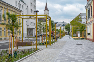 Städtische Straße mit gelben Gerüsten für städtische Begrünung, jungen Bäumen, Kopfsteinpflasterweg, Gebäuden und Radweg.
