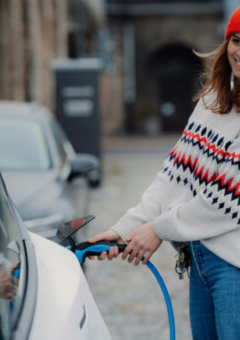 Eine junge Frau mit einem warmen Lächeln lädt ihr Elektroauto an einer öffentlichen Ladestation auf. Sie trägt einen gemütlichen, gemusterten Pullover, eine rote Mütze und Jeans, während sie den blauen Ladestecker anschließt. Die urbane Kulisse mit alten Gebäuden im Hintergrund vermittelt eine nachhaltige und moderne Atmosphäre.