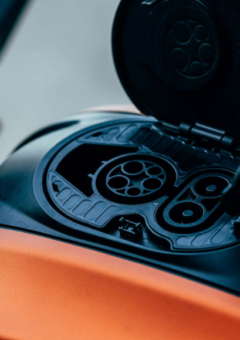 Nahaufnahme eines Ladeanschlusses an einem Elektromotorrad mit offenem Ladeport. Das Motorrad hat eine glänzende orangefarbene Verkleidung, und das Ladefach ist elegant in den Tankbereich integriert. Die modernen Steckverbindungen deuten auf eine leistungsstarke, zukunftsorientierte Mobilitätslösung hin.