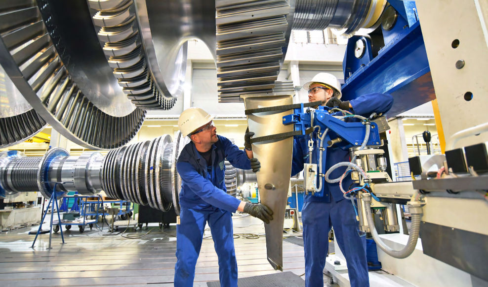 Zwei Techniker in blauer Arbeitskleidung montieren eine große Turbinenschaufel in einer Industrieanlage
