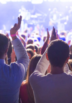 Eine Menschenmenge jubelt und applaudiert vor einer beleuchteten Bühne während eines Konzerts oder Events. Die Szene zeigt eine lebendige Atmosphäre mit Lichtern und verschwommenen Umrissen der Musiker im Hintergrund.