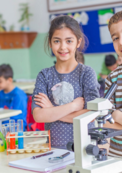 Zwei Kinder in einem Klassenzimmer stehen nebeneinander und lächeln in die Kamera, mit verschränkten Armen. Auf dem Tisch vor ihnen befinden sich ein Mikroskop, Reagenzgläser und eine kleine Pflanze. Im Hintergrund sind weitere Schüler und eine grüne Wandtafel zu sehen.