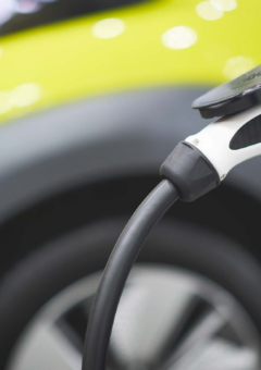 Ein Elektrofahrzeug wird an einer Ladestation aufgeladen. Im Vordergrund ist das Ladekabel in der Ladesteckdose zu sehen, während im Hintergrund verschwommen ein gelbes Auto erkennbar ist, das moderne und nachhaltige Mobilität symbolisiert.