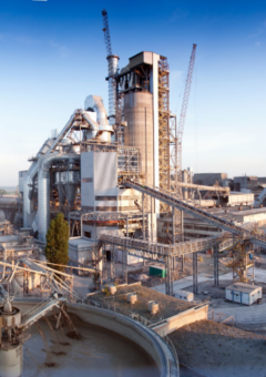 Eine industrielle Anlage mit großen Gebäuden, Türmen und Förderbändern, die sich durch das Gelände erstrecken. Im Vordergrund sind runde Wasserbecken zu sehen, während im Hintergrund Schornsteine und Kräne aufragen. Der Himmel ist klar und blau, was einen Kontrast zur industriellen Szene bietet.