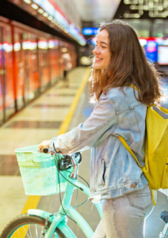 Eine junge Frau mit gelbem Rucksack und einem türkisfarbenen Fahrrad steht auf dem Bahnsteig einer U-Bahn-Station neben einem roten Zug. Die Atmosphäre ist hell und modern, mit reflektierenden Böden und leuchtenden Anzeigen im Hintergrund.