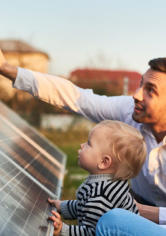 Eine Familie, bestehend aus einem Mann, einer Frau und einem kleinen Kind, sitzt neben einer Solaranlage im Freien. Der Mann zeigt lächelnd auf etwas in der Ferne, während das Kind neugierig auf die Solarmodule schaut. Die Szene vermittelt einen Eindruck von Nachhaltigkeit und Zukunftsdenken.