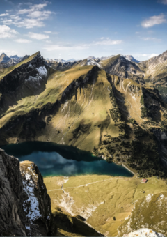 Panoramablick auf eine beeindruckende Berglandschaft mit schroffen Gipfeln, grünen Hängen und zwei klaren Bergseen. Die Szenerie ist von einem blauen Himmel mit vereinzelten Wolken gekrönt und vermittelt eine ruhige, majestätische Atmosphäre.