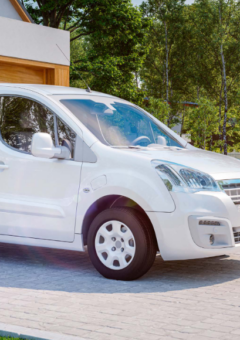 Ein weißer elektrischer Lieferwagen, der vor einer modernen Garage parkt und an einer Ladestation aufgeladen wird. Im Hintergrund sind grüne Bäume und ein gepflegter Garten zu sehen, die eine nachhaltige und umweltfreundliche Atmosphäre unterstreichen.