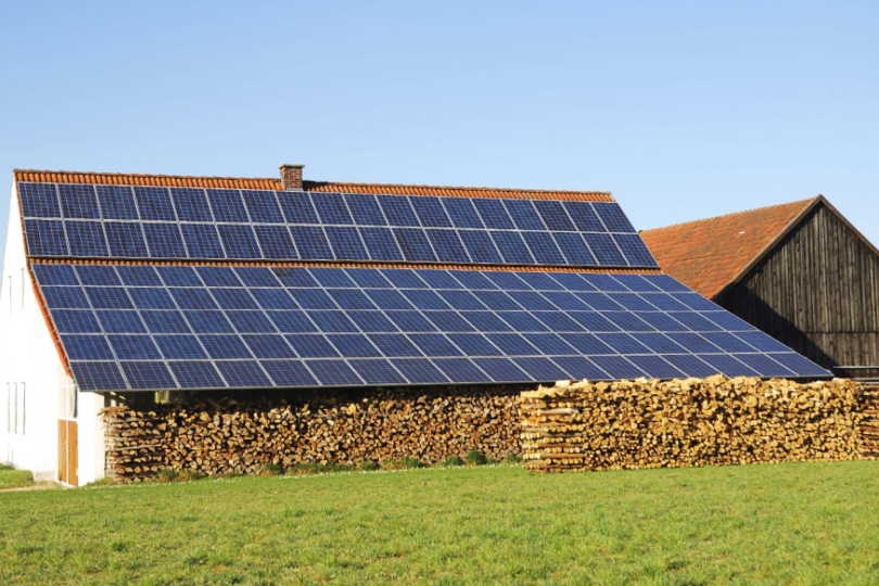 Ein landwirtschaftliches Gebäude mit einem Dach, das vollständig mit Solarmodulen bedeckt ist. Vor dem Gebäude ist gestapeltes Brennholz zu sehen, umgeben von einer grünen Wiese, und im Hintergrund befinden sich weitere Scheunen unter einem klaren blauen Himmel.
