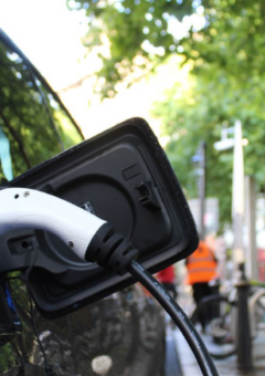 Ein schwarzes Elektroauto wird an einer Ladestation aufgeladen, mit einem weißen Ladekabel, das in den Ladeanschluss eingesteckt ist. Im Hintergrund sind grüne Bäume und eine städtische Straße zu sehen, die eine umweltfreundliche Mobilität symbolisieren.
