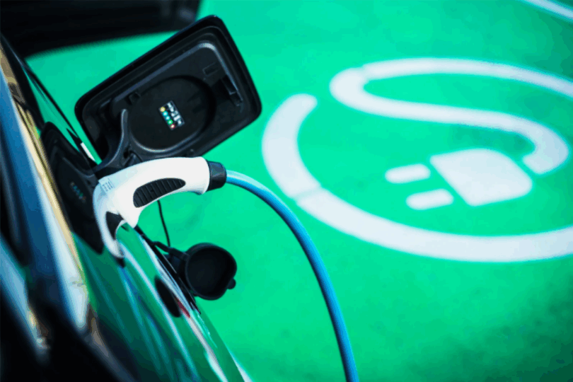 Nahaufnahme eines Elektroautos, das an einer Ladestation angeschlossen ist. Ein weißer Ladestecker mit blauem Kabel ist mit dem Fahrzeug verbunden. Im Hintergrund ist der grüne Boden mit einem weißen Symbol eines Steckers und Kabels zu sehen, das den Ladebereich für Elektrofahrzeuge kennzeichnet.
