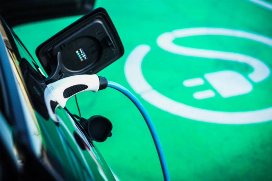 Nahaufnahme eines Elektroautos, das an einer Ladestation angeschlossen ist. Ein weißer Ladestecker mit blauem Kabel ist mit dem Fahrzeug verbunden. Im Hintergrund ist der grüne Boden mit einem weißen Symbol eines Steckers und Kabels zu sehen, das den Ladebereich für Elektrofahrzeuge kennzeichnet.
