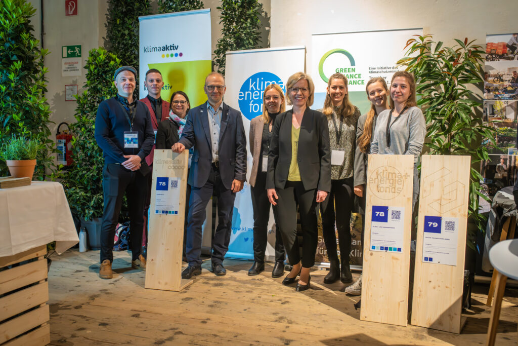 Eine Gruppe von acht Personen posiert für ein Foto auf einer Veranstaltung zum Thema Klimaschutz und Nachhaltigkeit. Sie stehen vor Bannern mit den Logos von klimaaktiv, Klima- und Energiefonds und Green Finance Alliance. Zwei der Personen halten große Holztafeln mit den Nummern 78 und 79, die mit QR-Codes und Projektinformationen versehen sind. Die Atmosphäre ist professionell, mit vielen Pflanzen im Hintergrund.