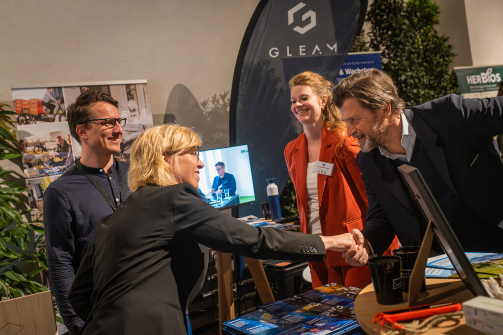 Eine Frau in einem schwarzen Blazer schüttelt die Hand eines Mannes in dunkler Kleidung an einem Messestand der Marke „GLEAM“. Eine Frau in einem orangefarbenen Blazer lächelt daneben, während ein Mann mit Brille im Hintergrund steht. Der Stand ist mit Broschüren, einem Bildschirm und Bannern ausgestattet, die das Unternehmen präsentieren.