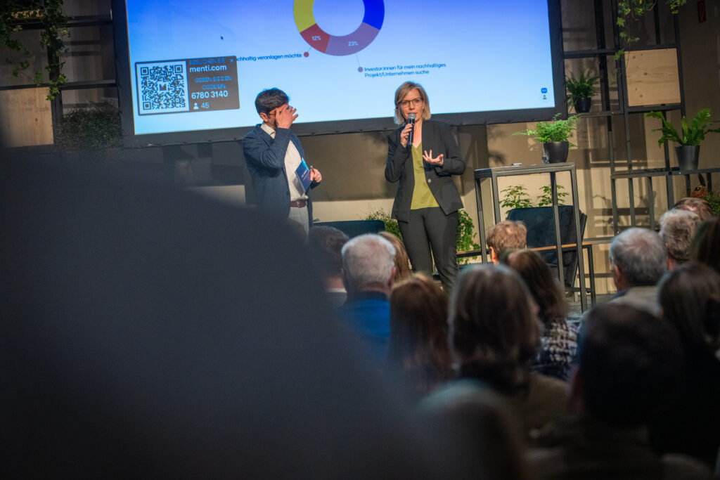 Eine Frau in einem schwarzen Blazer spricht mit einem Mikrofon vor einem Publikum, während ein Mann neben ihr mit einer Broschüre in der Hand sein Gesicht berührt. Hinter ihnen zeigt eine große Leinwand eine Präsentation mit einem Kreisdiagramm und einem QR-Code. Die Bühne ist mit Pflanzen dekoriert, und das Publikum folgt aufmerksam der Diskussion.