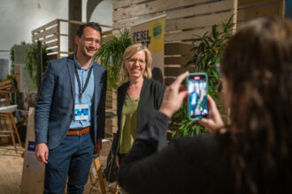 Ein Mann in einem blauen Blazer und eine Frau in einem schwarzen Blazer mit grünem Oberteil posieren lächelnd für ein Foto. Im Vordergrund hält eine Person ein Smartphone und nimmt das Bild auf. Im Hintergrund sind eine Holzverkleidung mit Pflanzen und ein Banner mit der Aufschrift „REBEL 100% BIO“ zu sehen, was auf ein nachhaltiges Thema hinweist.