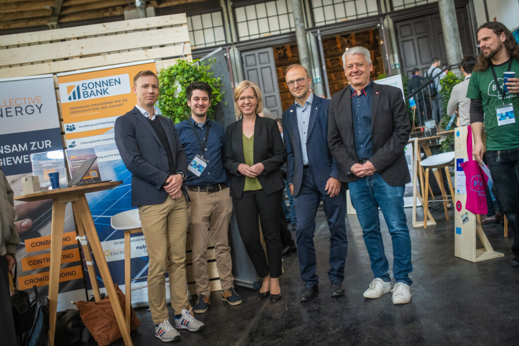 Eine Gruppe von fünf Personen steht zusammen und posiert für ein Foto auf einer Messe oder Konferenz. Im Hintergrund sind Stände mit Bannern zu sehen, darunter eines mit der Aufschrift „SONNEN BANK – Gemeinsam finanzieren wir die Energiesysteme der Zukunft“. Die Personen tragen Namensschilder, Business-Kleidung oder smarte Casual-Outfits, was auf ein Networking-Event oder eine Fachveranstaltung hindeutet.