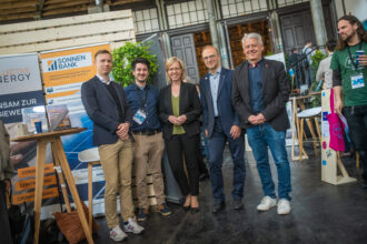 Eine Gruppe von fünf Personen steht zusammen und posiert für ein Foto auf einer Messe oder Konferenz. Im Hintergrund sind Stände mit Bannern zu sehen, darunter eines mit der Aufschrift „SONNEN BANK – Gemeinsam finanzieren wir die Energiesysteme der Zukunft“. Die Personen tragen Namensschilder, Business-Kleidung oder smarte Casual-Outfits, was auf ein Networking-Event oder eine Fachveranstaltung hindeutet.