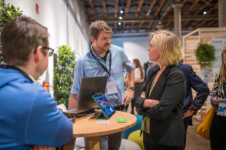 Ein Mann in einem hellblauen Hemd mit Namensschild spricht an einem Messestand mit einer Frau in einem schwarzen Blazer. Beide wirken konzentriert, während ein Tablet und ein Laptop auf einem runden Holztisch vor ihnen stehen. Im Hintergrund sind weitere Messebesucher und eine moderne, offene Veranstaltungsfläche mit Holz- und Pflanzenelementen zu sehen.