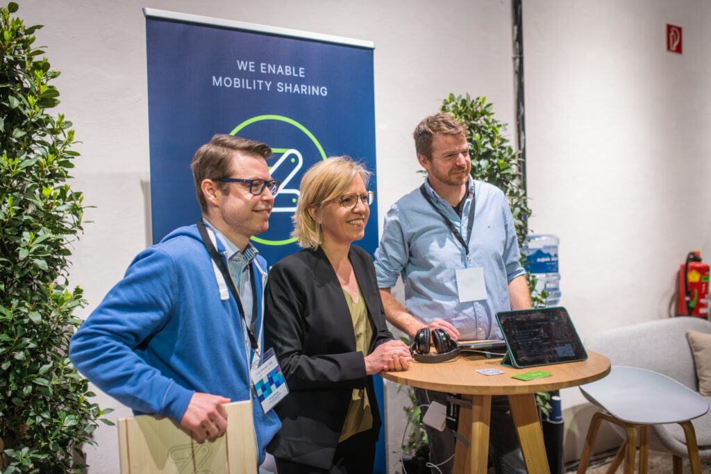 Drei Personen stehen vor einem Messe-Stand mit dem Banner „We Enable Mobility Sharing“ und lächeln in die Kamera. Einer der Männer trägt eine blaue Kapuzenjacke und hält eine hölzerne Auszeichnung, während die Frau in einem schwarzen Blazer neben ihm steht. Der dritte Mann, mit einem hellblauen Hemd und Namensschild, lehnt locker an einem runden Holztisch mit einem Tablet und Kopfhörern.