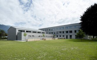Außenansicht eines modernen Gebäudekomplexes mit grauer Holzfassade und großen Fenstern, vor dem sich eine weitläufige Rasenfläche erstreckt. Mehrere Hochbeete aus hellem Holz mit Pflanzen sowie junge Bäume sind im Vordergrund platziert. Im Hintergrund sind bewaldete Berge zu sehen, während der Himmel teils bewölkt ist.