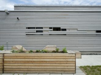 Ein rechteckiges Hochbeet aus hellem Holz, bepflanzt mit verschiedenen Kräutern und Gemüsepflanzen, steht vor einer grauen Holzfassade mit horizontalen Lamellen und kleinen Fenstern. Zwei integrierte Sitzbänke aus demselben Holzmaterial sind an den Enden des Hochbeets angebracht. Der Boden besteht aus Betonplatten und einem schmalen Rasenstreifen, während eine Außenleuchte an der Wand für Beleuchtung sorgt. Der Himmel im Hintergrund ist leicht bewölkt.